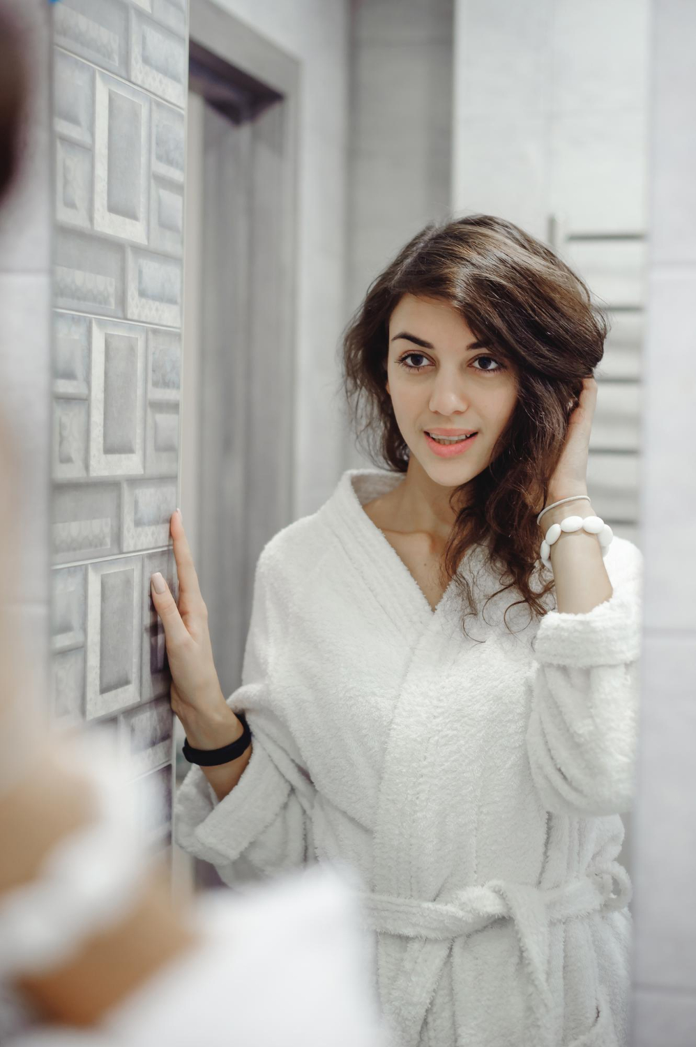 A woman with short brown hair smiling while touching her neck in front of a mirror, reflecting after her bariatric mental health evaluation at Advanced Client Care