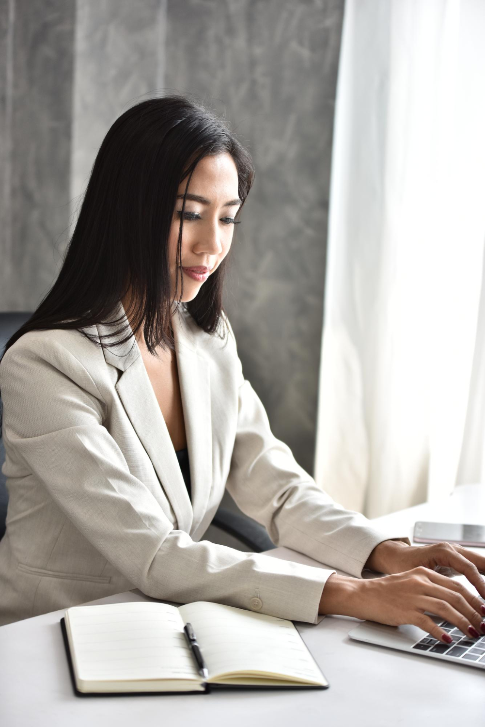 A Latina woman in a gray blazer writes on paper while sitting at a desk with a laptop, reflecting on stress management counseling techniques at Advanced Client Care